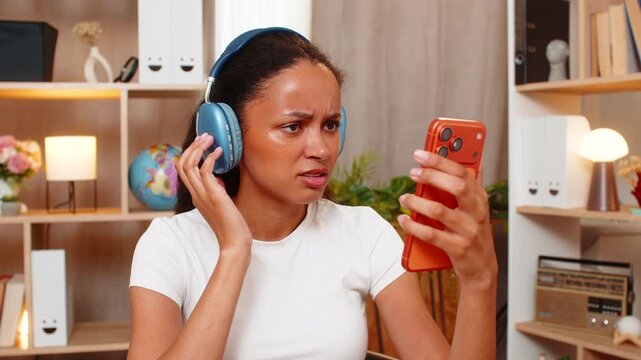 African American young woman on home table studies foreign language on smartphone and struggles to pronounce words correctly. Black girl tries speaking once more with focused face during home study.
