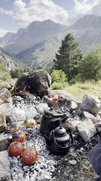 Vertical video of a hiker relaxing with stretched legs by a campfire roasting potatoes, onions, and peppers on glowing embers with a breathtaking alpine mountain peak landscape view