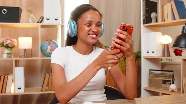 African American young woman on home table takes language test on smartphone and sees right answer. Black girl makes notes in notebook while studying words with headphones in an online lesson at home.