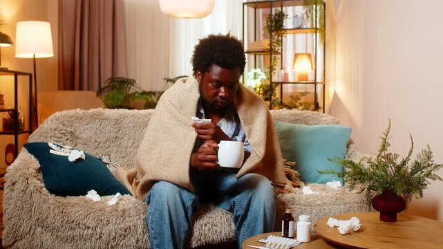 African American young man on home sofa sits under blanket holding warm tea and looking tired from illness. Black guy on home sofa keeps warm during cold recovery and rests with medicine after flu.