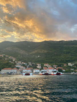 Several car ferries are docked at a coastal village harbor at the foot of a large green mountain during sunset