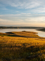 Obraz premium A vertical landscape capture of a glowing grassy meadow lit by warm golden hour sunlight, sloping down toward a serene lake surrounded by rolling hills and soft evening clouds.