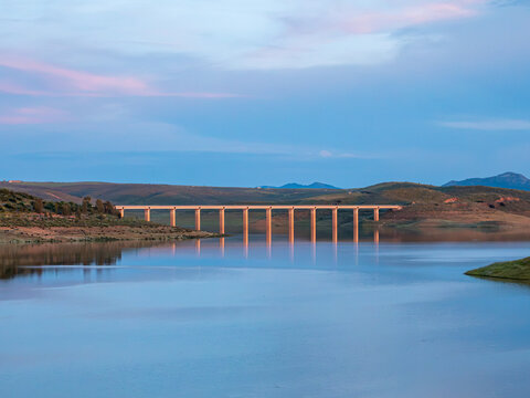 A wide horizontal shot of a long concrete bridge spanning across a peaceful reservoir, with its repeating pillars reflecting in the water under a soft twilight sky.