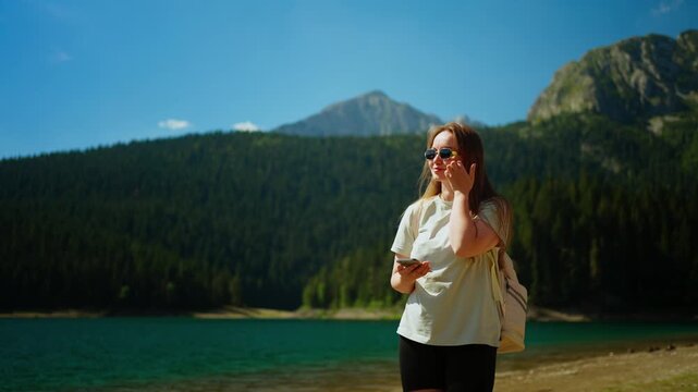 Young woman walking along a tranquil mountain lake shoreline, enjoying sunny weather, forest scenery, and peaceful nature during an outdoor summer getaway hike