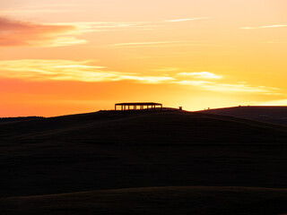 A striking silhouette of a simple roofed structure atop a dark rolling hill against a warm, glowing yellow and orange sunset sky. © Licia.Galli