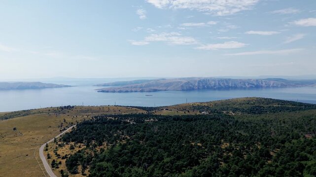 Aerial video of a broad coastal panorama with rugged mountain terrain, forested slopes and dry grasslands in the foreground. The calm Adriatic Sea stretches toward distant rocky islands