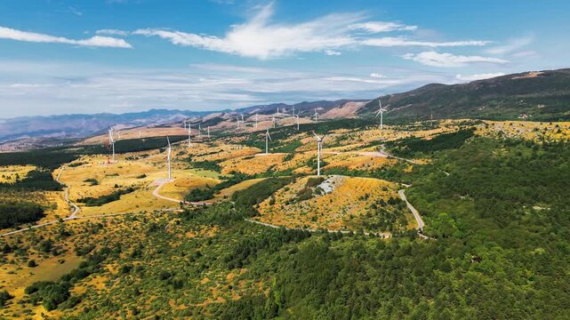 Aerial video over a vast mountainous landscape with numerous wind turbines distributed across golden grasslands and green forested hills