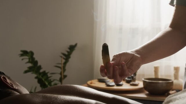 Caucasian masseuse holding smoking palo santo wood stick over back of African American woman during spa ritual. Useful for holistic wellness, body relaxation and massage therapy advertising