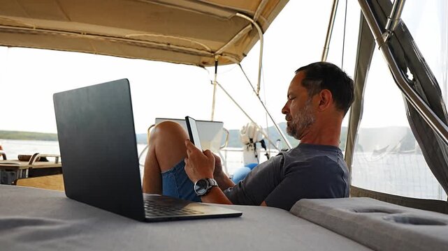 Relaxed man using laptop and smartphone while lounging on a sailing yacht. Digital nomad lifestyle and work life balance at sea.