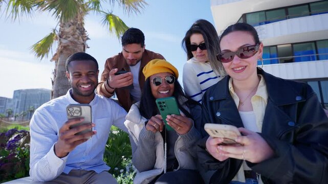 A seated group of five multiethnic young friends uses smartphones, compares screens and shares online content together.