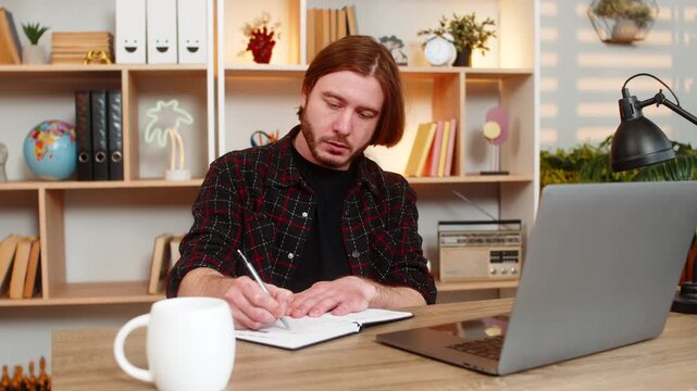 Young man on home table watches study video on laptop stops playback and writes ideas in notebook. Adult guy on home table stays focused on online learning and keeps taking notes with calm attention.