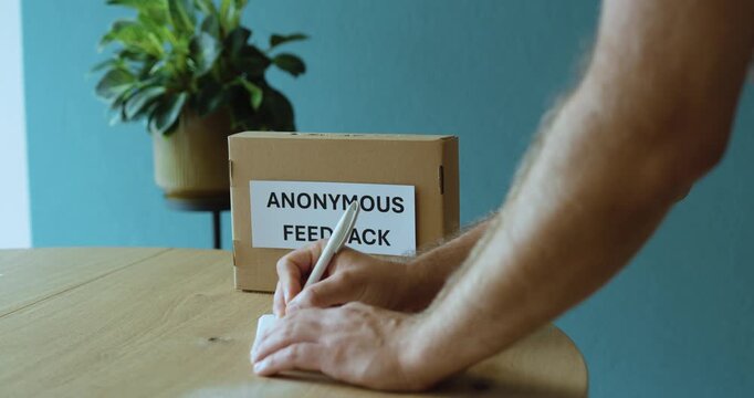 Man writing an anonymous feedback note for submission into a feedback box, representing confidential opinions, employee communication and workplace improvement.