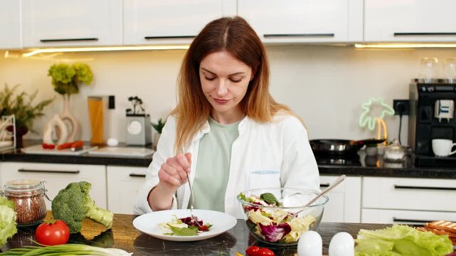 Middle-aged woman looks sad while eating vegetable salad in home kitchen for strict diet and weight loss daily goal. Young adult girl struggles with willpower discipline and healthy nutrition choice.