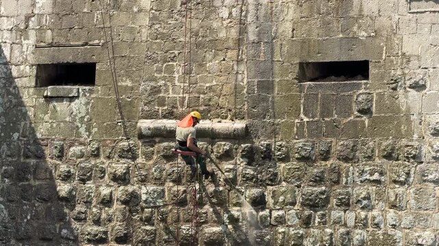 Maintenance worker suspended on ropes cleans an ancient stone fortress wall with a high-pressure washer