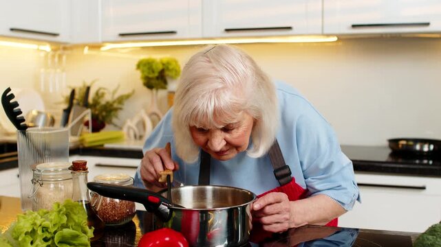 Senior old woman stirs soup in home kitchen smells aroma then tastes broth feeling satisfied for healthy diet nutrition. Elderly grandmother enjoys warm meal smiling during indoor lifestyle routine.
