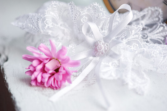 White lace wedding garter tied with a delicate ribbon and a pink daisy-style flower on a worn wooden surface, evoking romantic, vintage bridal charm and soft, feminine detail for celebrations.