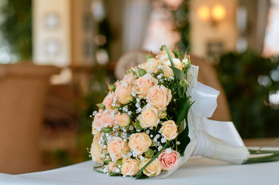 Peach and cream roses arranged with baby's breath and greenery, tied with a white ribbon. Soft romantic wedding bouquet styled on a table in a warm, elegant indoor reception setting.