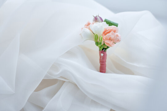 Delicate peach and white boutonniere rests on soft flowing white fabric, evoking wedding elegance, romance and bridal details; a small wrapped floral accessory ideal for ceremonies and portraits.