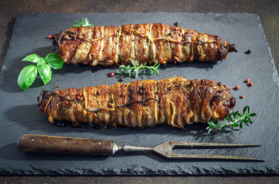 Two roasted pork necks wrapped in bacon on slate platter