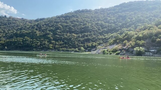 Two double kayaks paddle across a green, scenic river bordered by a lush forested mountain