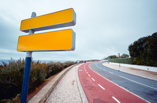 Blank yellow signpost beside a seaside promenade and red bike lane, overlooking a curving coastal road with pedestrians under an overcast sky &mdash; a quiet, minimal travel and wayfinding scene.