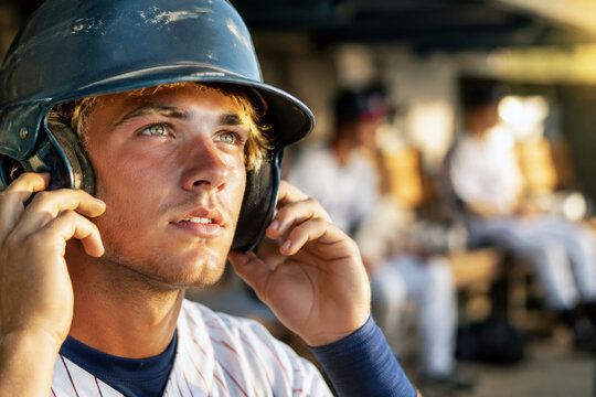 A young baseball player adjusts his dark batting helmet with both hands while looking upward from a sunlit dugout. Focused preparation