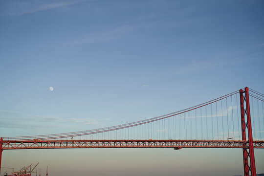 A red suspension bridge spans across the image with a clear blue sky and visible moon.