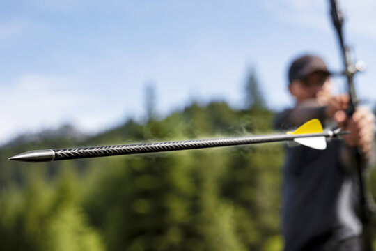 A sharp arrow flies across a blurred natural background while its fletching and textured shaft remain crisp in sunlight. Speed and accuracy