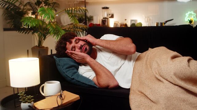 Arabian man at home lies still with heavy headache holds head at temple closes eyes breath slow. Egyptian guy on sofa takes pill drinks water dims screen stops sound waits for relief stays quiet pain.