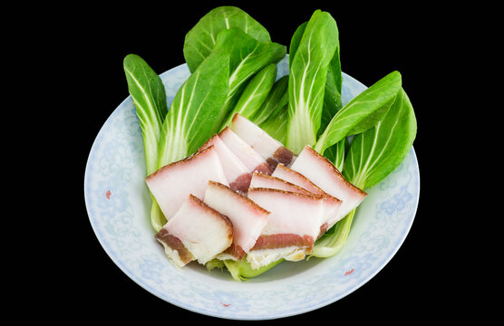 Sliced fatty pork belly rests on fresh bok choy, served on a light blue plate against a black background, raw food ingredients for cooking.