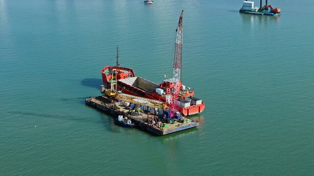 Crane barge and dredging vessel positioned side by side during seabed sediment removal. Industrial marine scene in calm coastal water
