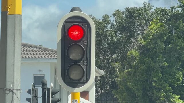 Close-up view of a traffic light showing red, mounted on a pole with a suburban home and lush green trees in the soft-focus background. Clear daytime lighting.