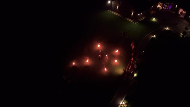 Drone video captures skilled fire dancers performing for a group of event guests on a lit lawn at night, with flaming props creating bright patterns and dramatic ambiance for the celebration.