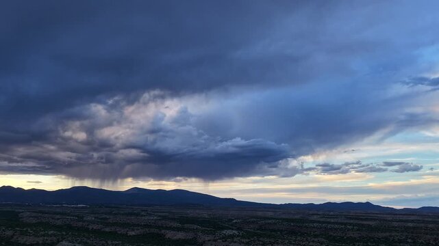 4K drone weather time lapse capturing a dramatic storm system over a southwestern landscape. Heavy rain clouds sweep across distant mountains and mesas as changing sunset light colors the sky.