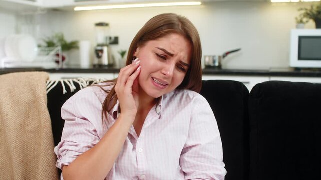 Young woman at home holds cheek with toothache and presses palm to jaw while eyes squint with pain. Caucasian girl on sofa breathes slow rubs gum waits for care and plans visit to dentist for relief
