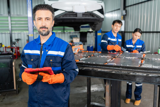 Bearded technician with a tablet faces the camera in an EV repair garage while colleagues test a battery pack behind him.