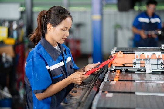 Beside an electric vehicle battery pack, a female technician uses a tablet while a male colleague stands out of focus behind.