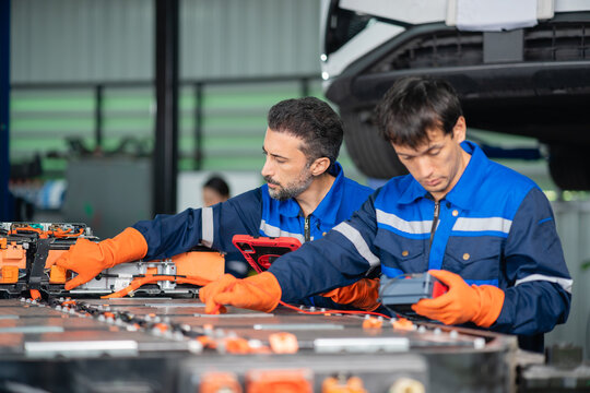 Beside an electric vehicle battery pack, a technician points out a module while his colleague holds an insulation tester ready.