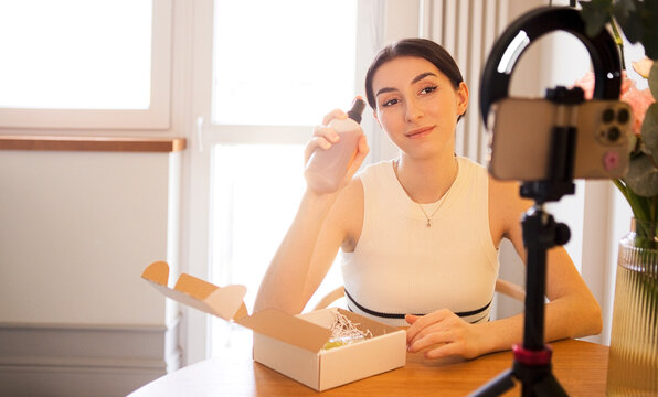 Young woman unboxing skincare product while recording video for social media, sitting at wooden table with smartphone on tripod and decorative flowers in background