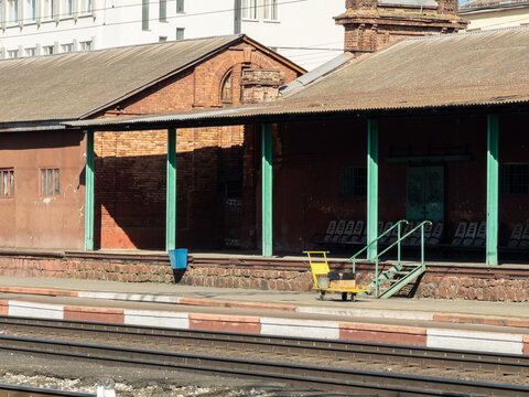An old brick train station platform with metal seats and a luggage trolley, portraying a nostalgic atmosphere of historical travel infrastructure in a quiet rural transport terminal