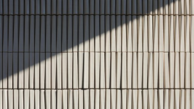 Close-up of a glazed ceramic fin rainscreen facade with rows of slender vertical tiles shifting from dark to cream under a diagonal shadow line. Contemporary architectural cladding material
