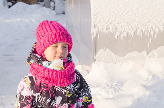 Happy little girl smiling and squinting in bright winter sun outdoors. Cute child in vivid pink knitted hat and colorful warm outerwear. Childhood, winter lifestyle and kids fashion concept,copy space