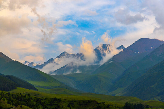 Breathtaking view of a mountain range with lush green foothills and imposing snow-dusted summits. Soft clouds drift across the sky, enhancing the scenic beauty of the landscape.
