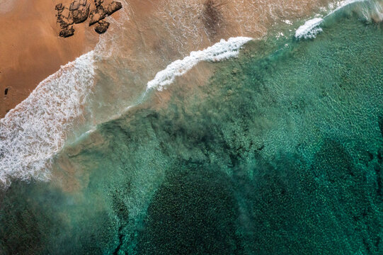 An ocean wave breaks along a turquoise coloured, pristine, reef-lined sandy shore. Captured on a beautiful idyllic sunny day from high above the beach on the South Coast of New South Wales, Australia.