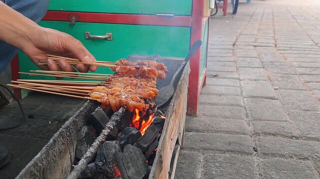 Hand grilling chicken satay skewers over charcoal fire. Indonesian street food, satay, barbecue, cooking, smoke, fire, UMKM, traditional, delicious.