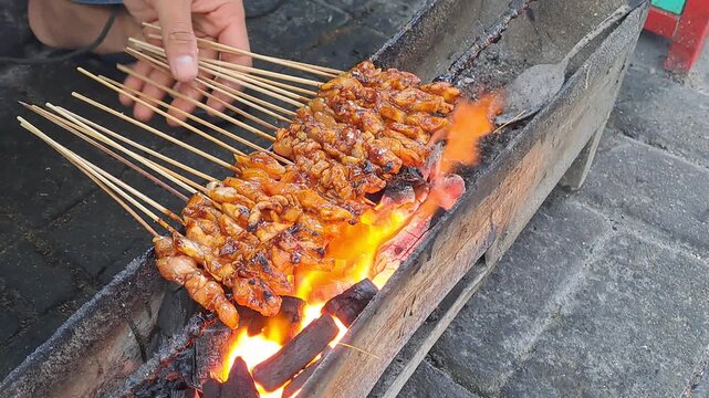 Hand grilling chicken satay skewers over charcoal fire. Indonesian street food, satay, barbecue, cooking, smoke, fire, UMKM, traditional, delicious.