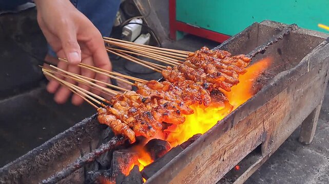 Hand grilling chicken satay skewers over charcoal fire. Indonesian street food, satay, barbecue, cooking, smoke, fire, UMKM, traditional, delicious.