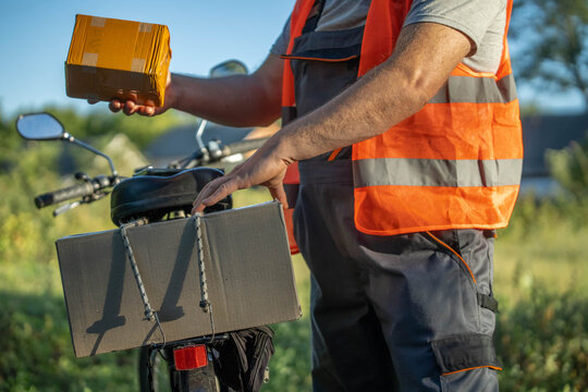 A delivery worker wearing a bright orange vest holds a small package while placing another in a cargo box on his bike. The sun shines in a lush, green area