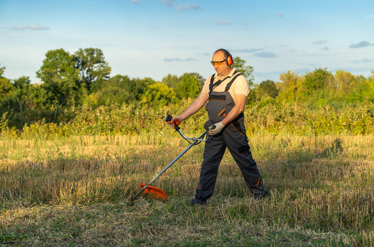 Farmer trims field grass with brush cutter in open meadow, wearing ear protection and work overalls. Concentrated worker handles tool with steady posture against tall trees and clear sky.
