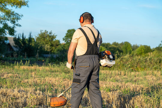 Male landscaper operates brush cutter in grassy field. Grounds maintenance worker wears overalls, gloves and hearing protection, standing amid dry stubble with tool at rest in warm evening light.